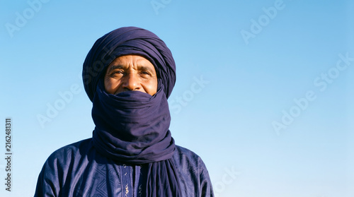 A close up portrait of a smiling Tuareg man wearing a traditional indigo blue tagelmust turban and robe, standing under a clear desert sky with a peaceful expression.