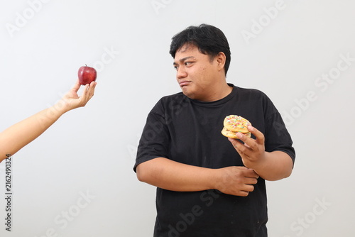 A heavyset man with a sad expression, holding a doughnut and glancing sideways at the hand of someone offering an apple, appears torn between the food choices.