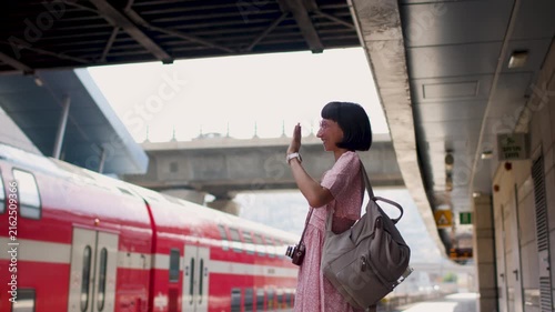 The girl in a pink dress and with a camera stands on the platform of the train station and waves with her hand. goodbye to friends. travel and vacation. creating content about travel. tourist with cam