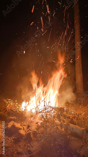 A bonfire blazes with large orange yellow flames reaching upward as sparks and embers fly into the dark night sky. Burning branches and foliage form the bright base, illuminating rocky ground.