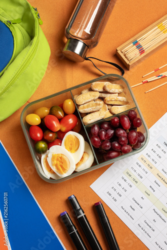 Healthy school lunchbox with boiled eggs, cherry tomatoes, grapes and crackers next to weekly class schedule, markers and backpack. Balanced kids nutrition and back to school concept.