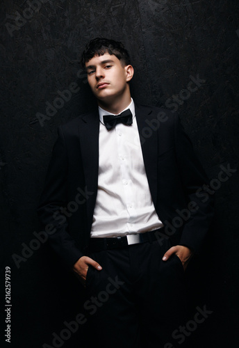 Formal elegance and luxury lifestyle, young man in a black tuxedo and bow tie posing with confidence against a dark textured wall for evening events