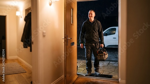 A smiling maintenance engineer with a tool bag stands at an open front door on a rainy night.