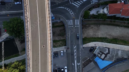 Bridge spans roadway.
Cars cross bridge.
Vehicles navigate intersection.
Houses surround streets.