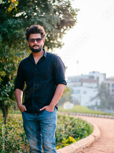 Canvas Print Young Indian man in sunglasses, casual attire, standing outdoors in a park, shallow depth of field