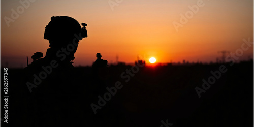 Silhouette of soldiers in tactical gear standing against a dramatic sunset sky background.