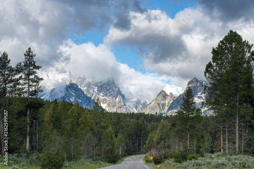 Scenic road winds through a dense pine forest toward the snowcapped Teton Range beneath dramatic low clouds in Grand Teton National Park, Wyoming, USA