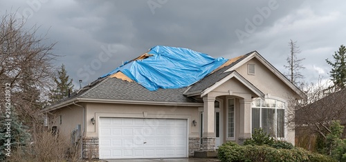A suburban house with a makeshift roof cover showing exterior damage under dark clouds, highlighting urgent repair needs.