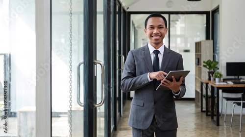 Busy young Indonesian business man executive standing in office using digital tablet. Young professional businessman corporate manager wearing suit and glasses holding tab working on finance project