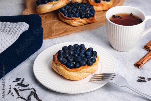 Blueberry puff pastry tart with fork on white plate, tea cup, cinnamon sticks and loose tea leaves on textured table, cozy breakfast scene