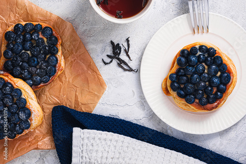 Blueberry puff pastry tart with fork on white plate, tea cup, cinnamon sticks and loose tea leaves on textured table, cozy breakfast scene, top view
