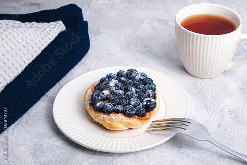 Blueberry puff pastry tart with sugar powder and fork on white plate, tea cup on textured table, cozy breakfast scene