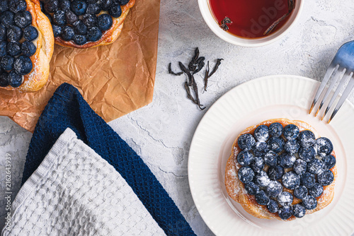 Blueberry puff pastry tart with sugar powder and fork on white plate, tea cup, cinnamon sticks and loose tea leaves on textured table, cozy breakfast scene, top view