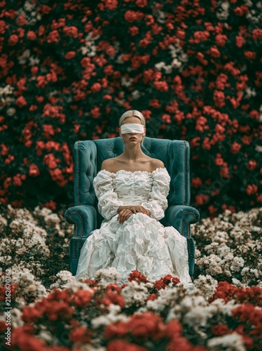 A young woman sits in an English armchair against a backdrop of flowers, her eyes hidden by a blindfold 
