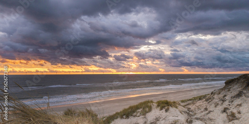 Aerial view of the sandy coastline with dunes, marram grass, and a dramatic sunset over the North Sea with distant wind turbines on the horizon in Hvide Sande, Denmark.