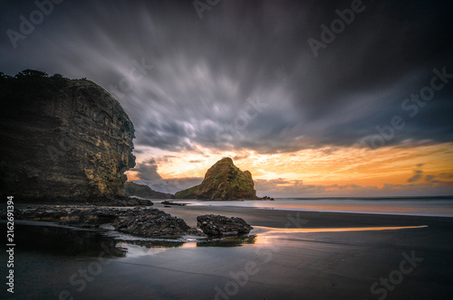 New Zealand - 15 April 2021: View of a dramatic sunset over a dark sand beach with a prominent rock formation and moody storm clouds.