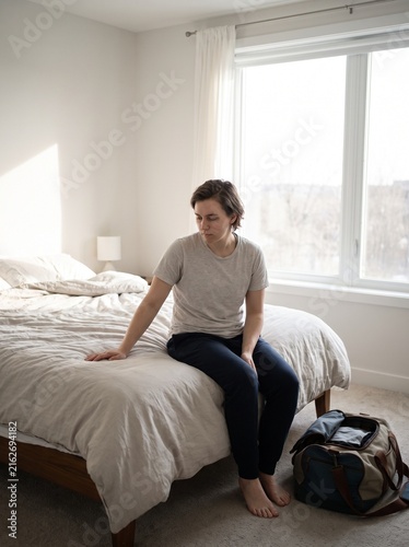 Thoughtful young adult sitting on the edge of a bed with a travel bag in a bright modern bedroom