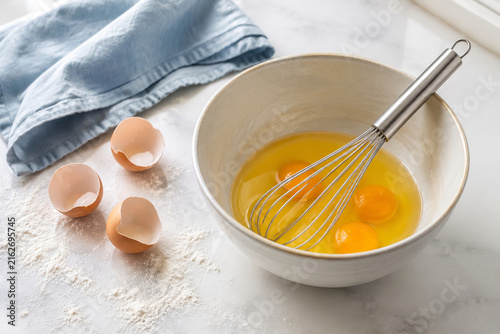 Stainless steel whisk in ceramic bowl with cracked eggs and flour on counter, baking preparation concept