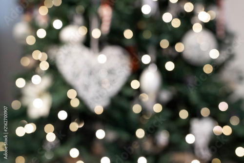Blurred close-up of a decorated Christmas tree with glowing lights, white heart ornament, and festive baubles bokeh background. The concept of the New Year and Christmas time.