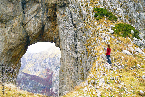 Fotografie Female adventurer standing beside massive natural limestone arch framing distant mountain valley in Prokletije