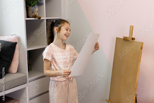 A cute young girl is looking at her artwork on a piece of paper after painting at home. Creative child admiring her drawing, standing near an easel in her room. Children's leisure concept