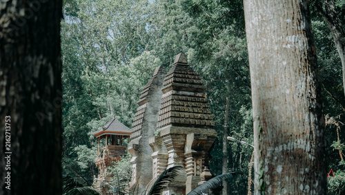 Cuadro en lienzo A picturesque view of the Candi Bentar split gate and a traditional Balinese Meru tower, partially obscured by a lush forest canopy and framed by large tree trunks in the foreground