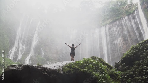 Young woman standing on a rock with arms outstretched, celebrating freedom at a powerful waterfall