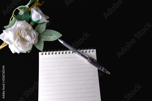 Blank spiral notebook pen and artificial white rose on black background