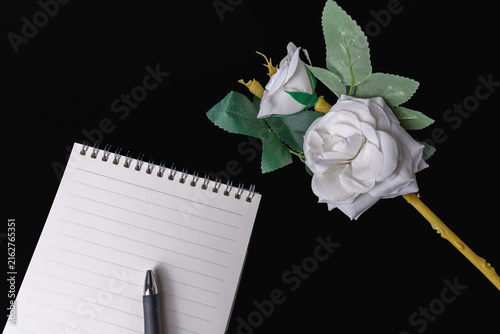 Blank spiral notebook pen and artificial white rose on black background
