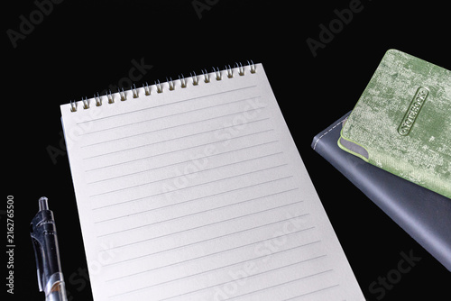 Blank lined spiral notepad pen and stacked closed notebooks on black background