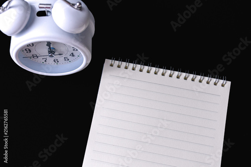White twin bell alarm clock and blank lined spiral notepad on black background