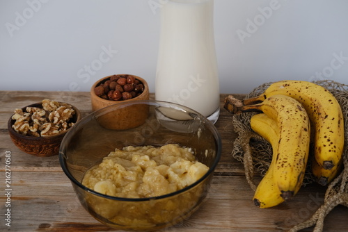 Slicing ripe bananas on wooden cutting board for healthy breakfast with fresh natural ingredients, glass milk and bowl of hazelnuts on rustic table, cooking process and nutritious morning meal concept
