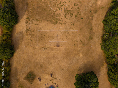 Aerial view of a dry, parched soccer field with white markings surrounded by lush green trees in Stafford, England, United Kingdom.