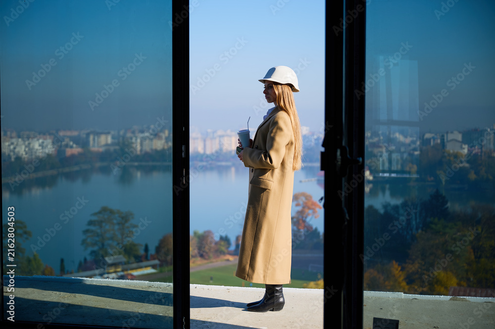 Fototapeta premium Female property manager standing on high rise terrace with coffee, showing scenic city and lake view. Concept of housing presentation, property development, urban living and business use