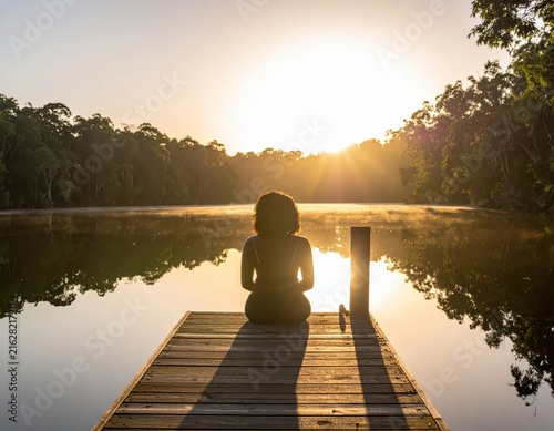 Serene Woman Sitting on Dock Enjoying Sunrise Reflections Over Calm Lake Surrounded by Nature