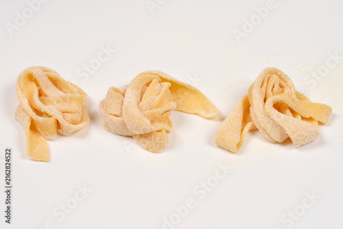 Three small nests of fresh raw homemade tagliatelle pasta lightly dusted with flour on a plain white surface.