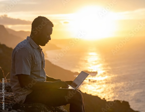 Man Working on Laptop at Sunset with Ocean View and Mountains in Background