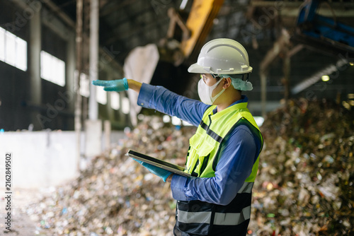 Environmental engineer in protective safety gear inspecting processed waste and taking notes on a clipboard at an industrial recycling facility.