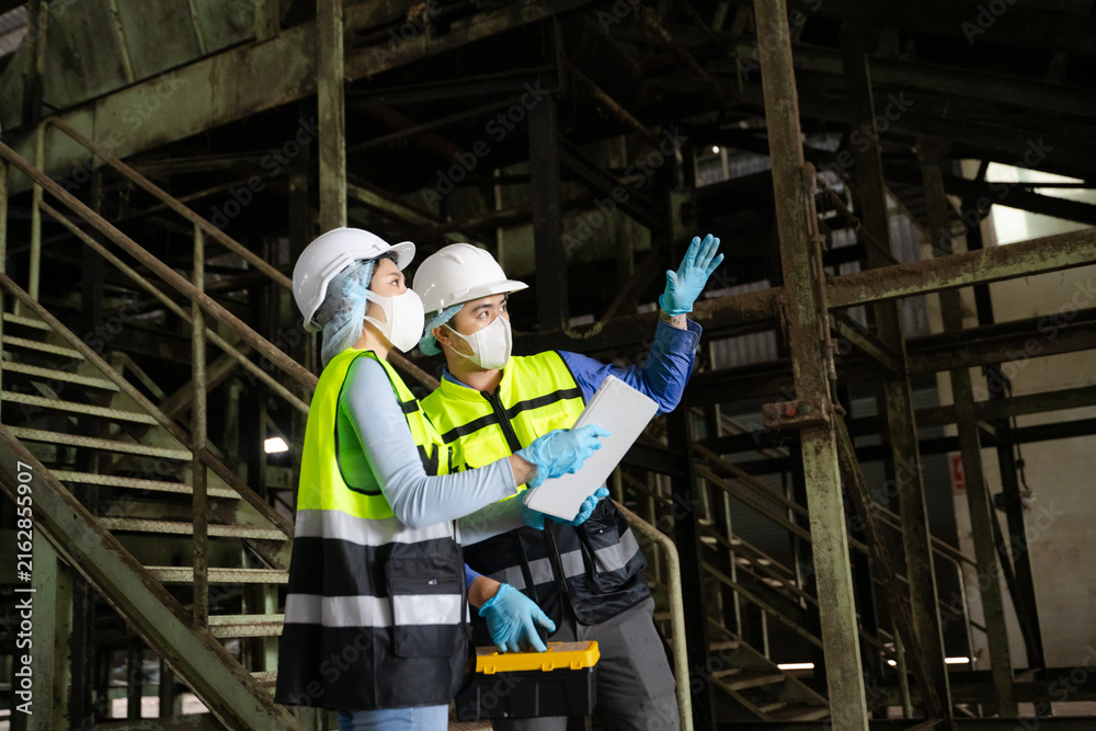 Fototapeta premium Professional male and female engineers wearing hardhats and masks inspect an industrial factory workflow using a digital tablet