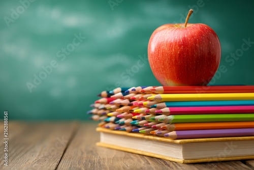 Colorful pencils and red apple on stacked books atop wooden desk with green background ideal for educational themes