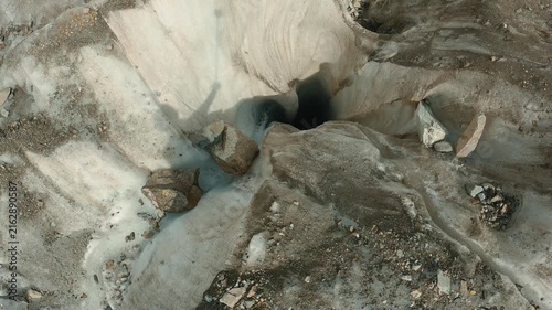 Aerial view of glacier ice cave with dark entrance, surrounded by rocky debris flowing meltwater. Soft natural lighting highlights icy textures gray white tones