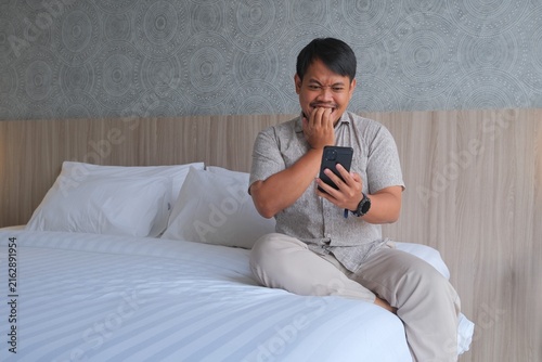 Fotografie Shocked Asian man sitting on a bed, holding a smartphone and touching his head while reacting with surprise to something on the screen