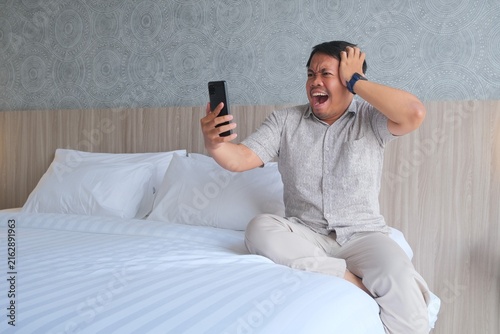 Принти на полотні Shocked Asian man sitting on a bed, holding a smartphone and touching his head while reacting with surprise to something on the screen