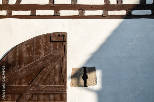 Hölzernes Rundbogentor an historischem Gebäude Zehntscheuer mit weiß gekalkter Wand mit Schießscharte und Fachwerk und Schatten eines Hausdaches mit Regenrinne in Neckartenzlingen.