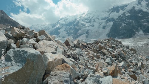 Timelapse of fast moving clouds over snowy mountains. Rugged boulders in foreground lead to vast glacier snow capped peaks under dramatic clouds. Cool blue gray tones dominate.