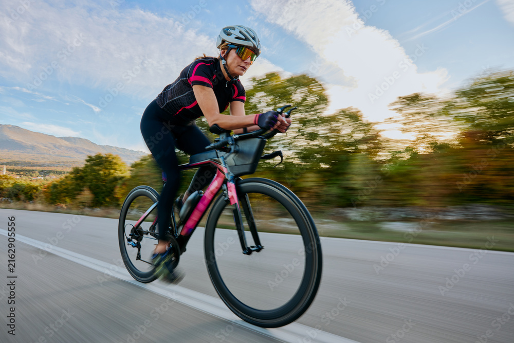 Naklejka premium Female Cyclist Racing on Road Bike at Speed Through Countryside During Sunset Training Ride