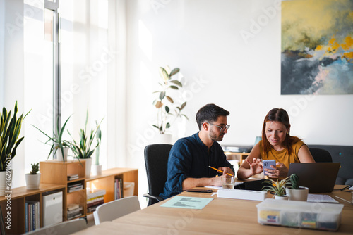 A male and a female colleague are collaborating in a bright modern office. They sit together at a desk, reviewing data on a smartphone while referencing a laptop.