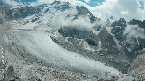 Timelapse clouds over snowy mountains. Glacier flows down rocky valley between jagged snowcapped peaks under partly cloudy sky. Mist rises from ice. Composition emphasizes scale raw natural power