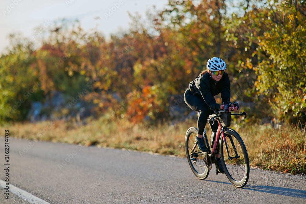 Fototapeta premium Female Cyclist Training on a Road Bike in Autumn Countryside Wearing Helmet and Sunglasses