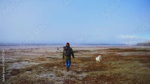 Person walks alone across vast, frosty field with dog. White dog trots beside person, exploring terrain. Clear sky, cold morning light casts long shadows. Scene evokes solitude, nature